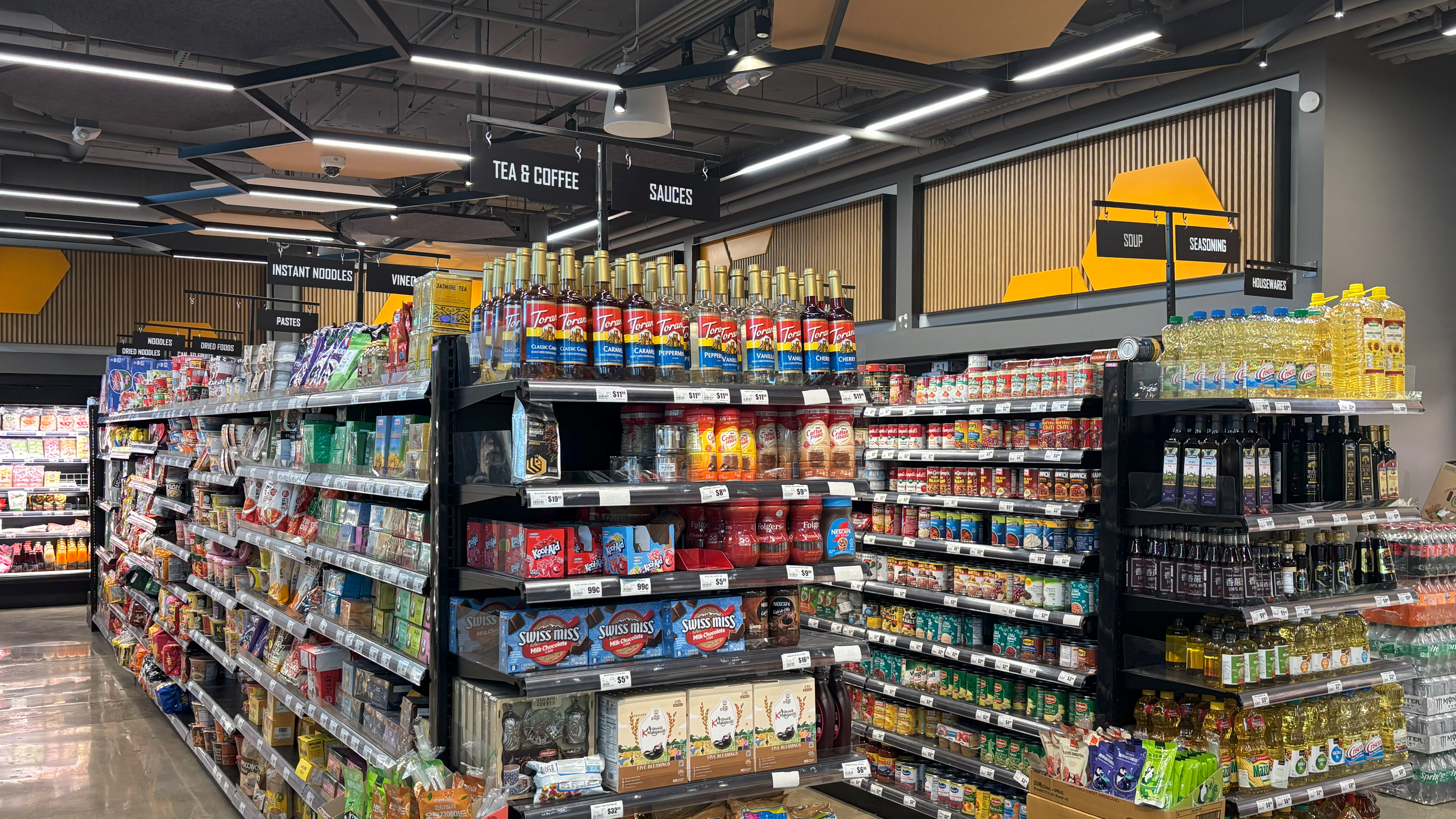 Aisle view at Yellow Bee Market & Cafe in Seattle featuring black gondola shelving, bold aisle signs for tea, coffee, sauces, and neatly stocked groceries.