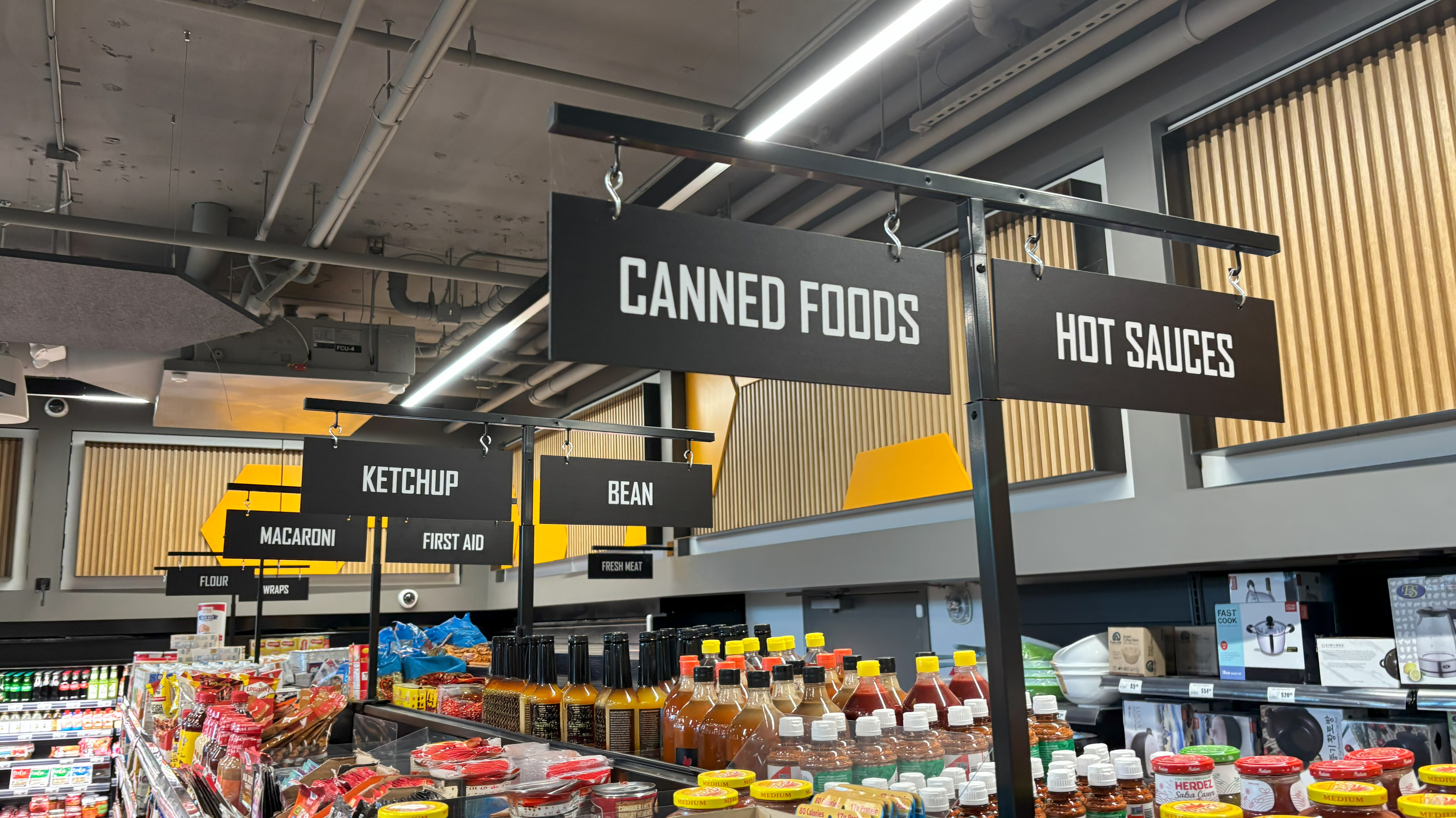 Black metal aisle sign holders with bold white category signs at Yellow Bee Market & Cafe in Seattle, helping shoppers navigate the compact supermarket.